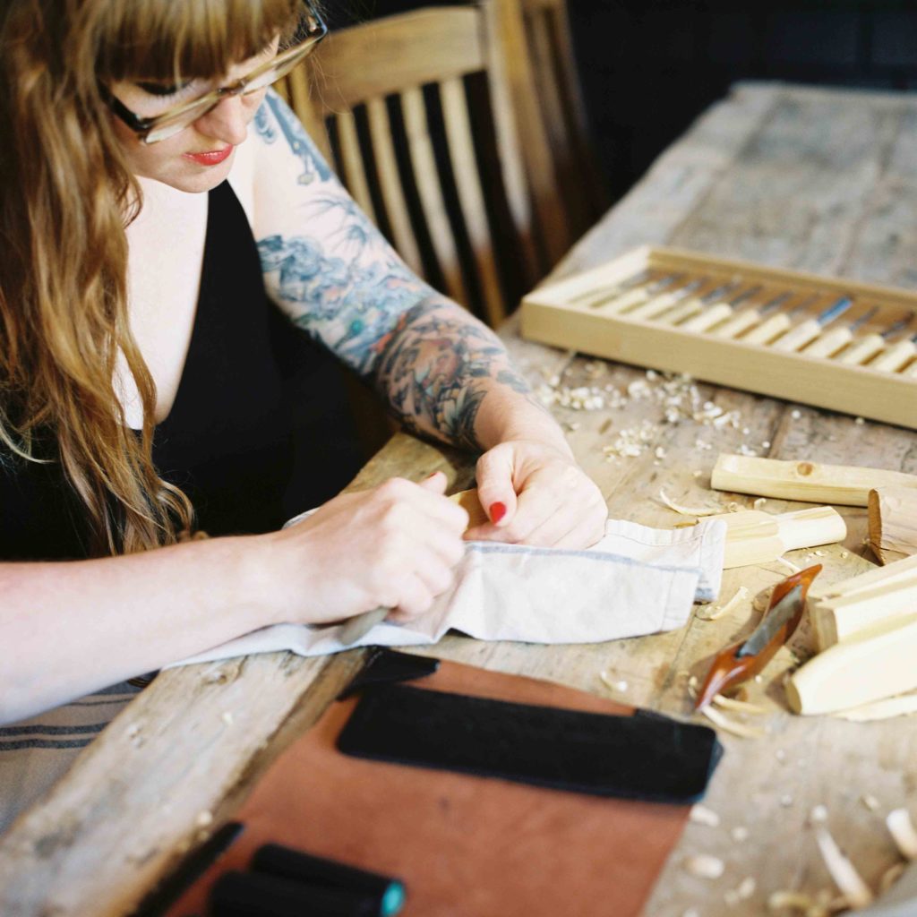 A woman with long hair and tattoos carves a piece of wood 